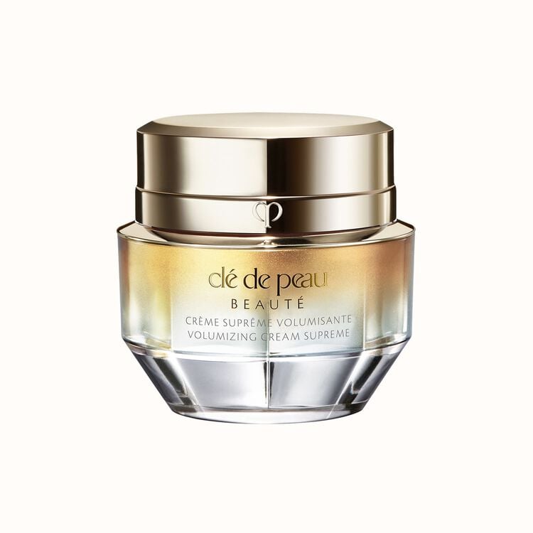 A jar of Clé de Peau Beauté Crème Suprême Volumisante Volumizing Cream Supreme. The jar has a gold, reflective lid and an ombré glass base that transitions from gold to clear.