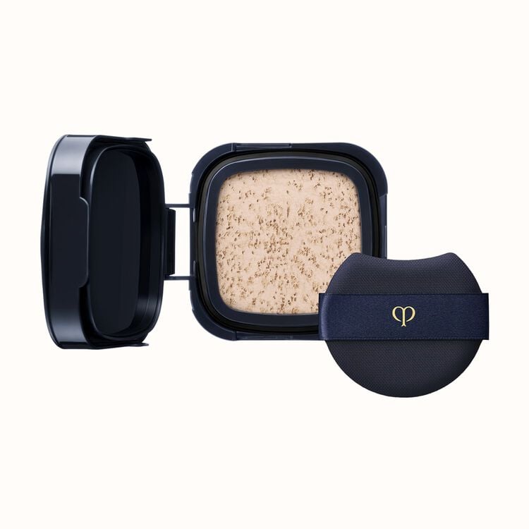 A black compact with an open lid revealing light-colored pressed powder, next to a black cushion puff with a gold logo on a dark blue ribbon.