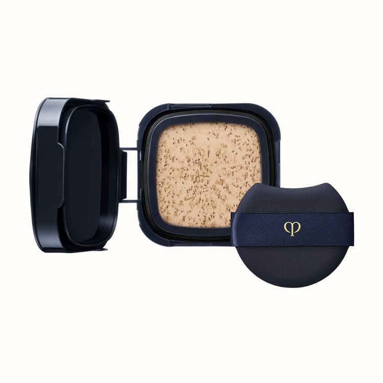 A black compact with an open lid revealing light-colored pressed powder, next to a black cushion puff with a gold logo on a dark blue ribbon.