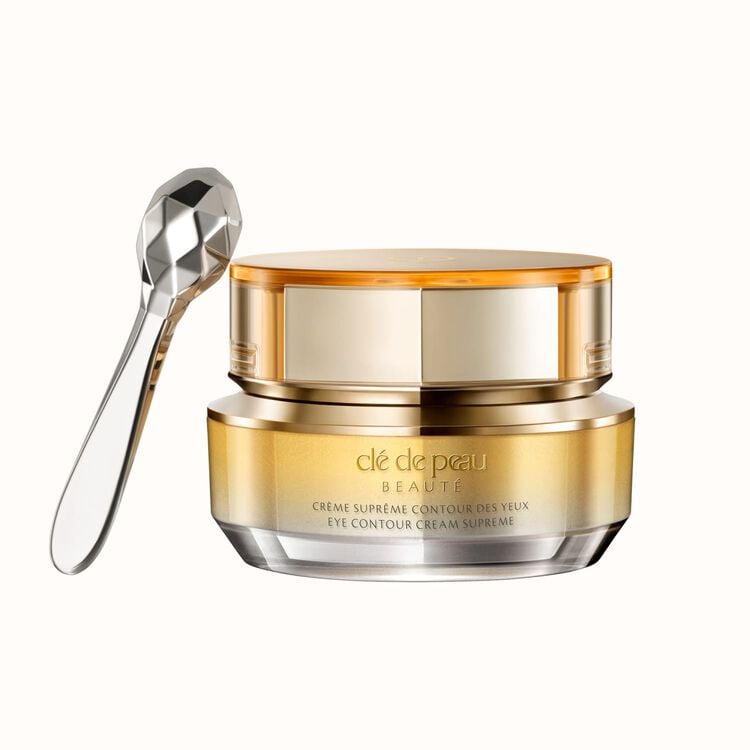 A luxurious gold and glass jar of Clé de Peau Beauté Eye Contour Cream Supreme with a silver, faceted applicator spoon resting against it, all on a white background.
