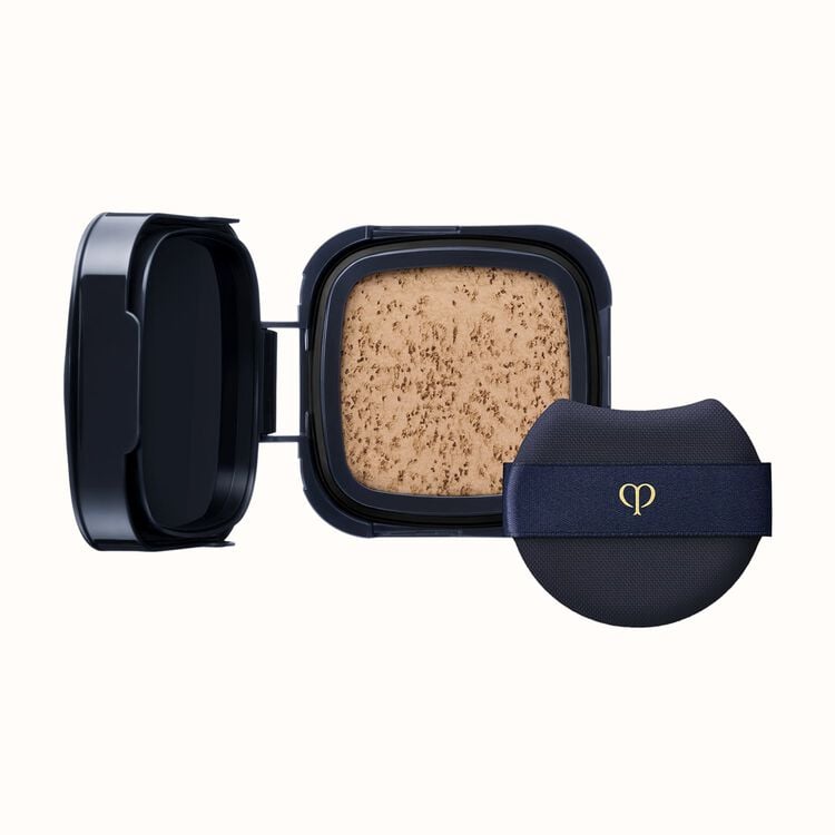 A black compact with an open lid revealing light-colored pressed powder, next to a black cushion puff with a gold logo on a dark blue ribbon.
