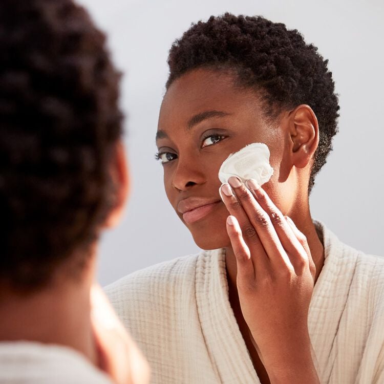 A woman with short, curly dark hair looks into a mirror, applying a white cream to her cheek with a cotton pad. She wears a light-colored, textured robe.