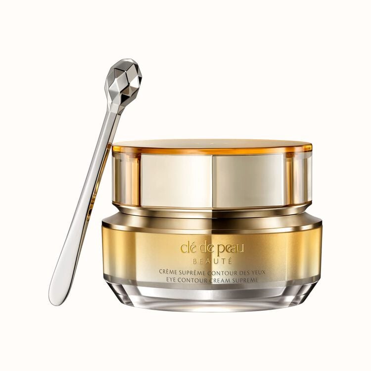 A luxurious gold and glass jar of Clé de Peau Beauté Eye Contour Cream Supreme with a silver, faceted applicator spoon resting against it, all on a white background.