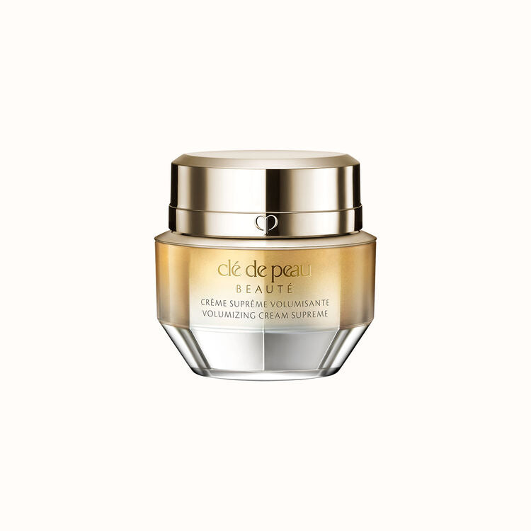 A jar of Clé de Peau Beauté Crème Suprême Volumisante Volumizing Cream Supreme. The jar has a gold, reflective lid and an ombré glass base that transitions from gold to clear.