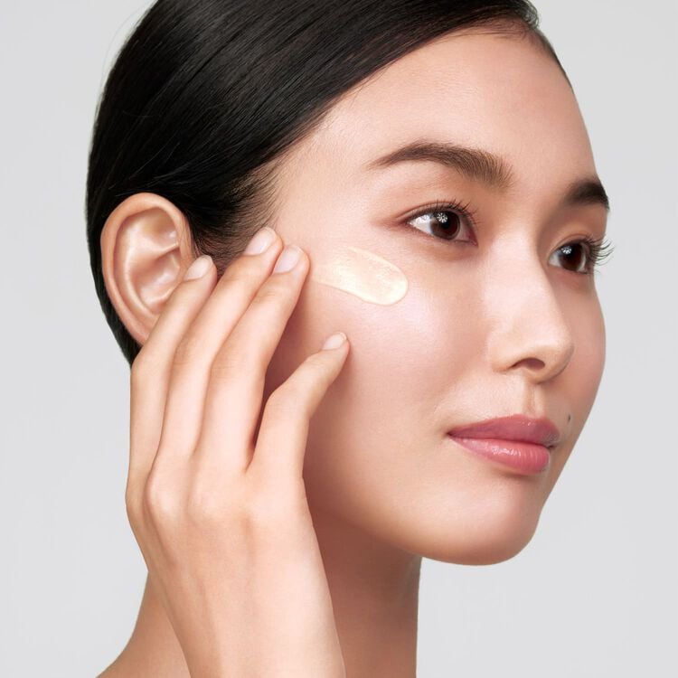 A woman with dark hair applying a streak of light-colored foundation or concealer to her cheek with her fingertips.