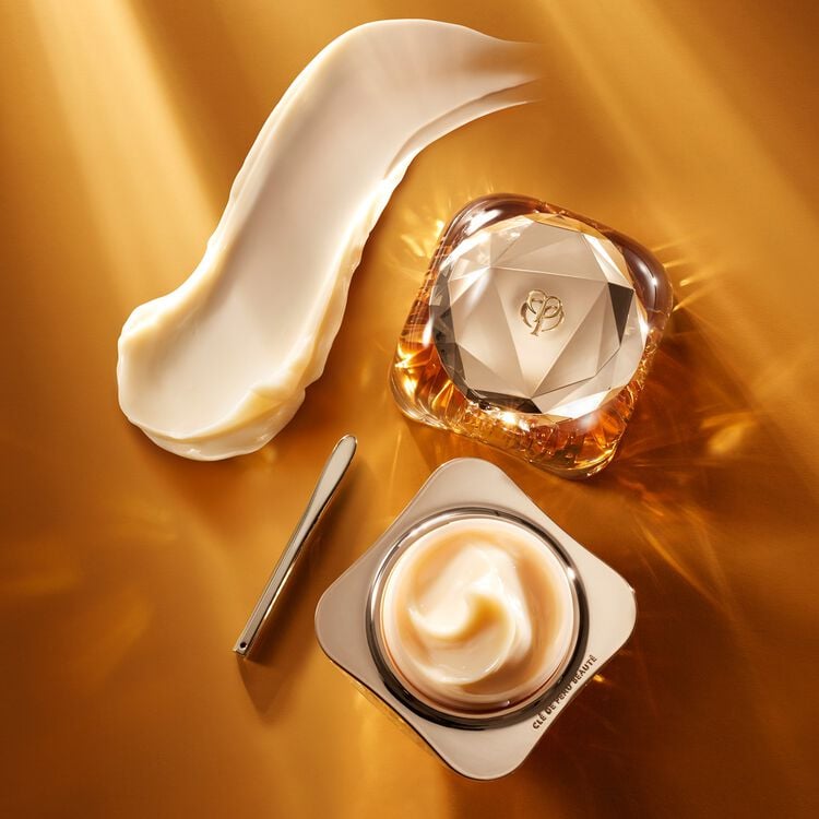 A luxurious gold and diamond-shaped jar of face cream, an open jar of the same cream with a dollop of product, a silver spatula, and a large swipe of the cream on a golden surface.