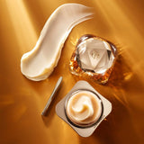 A luxurious gold and diamond-shaped jar of face cream, an open jar of the same cream with a dollop of product, a silver spatula, and a large swipe of the cream on a golden surface.
