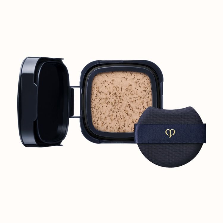 A black compact with an open lid revealing light-colored pressed powder, next to a black cushion puff with a gold logo on a dark blue ribbon.