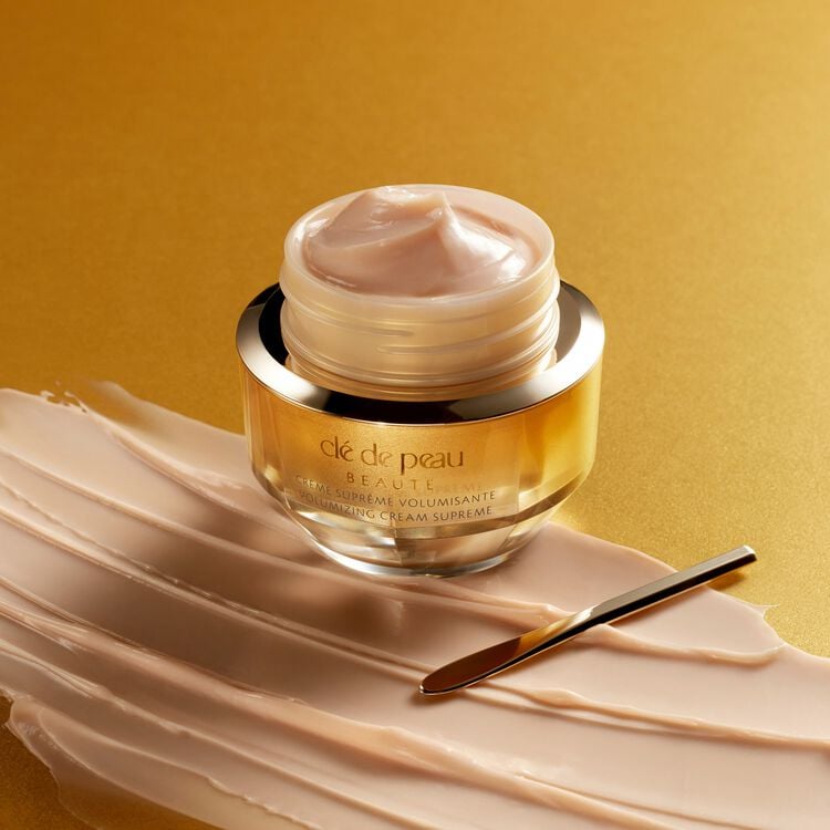 A golden jar of Clé de Peau Beauté Volumizing Cream Supreme, with a swirl of the pink-toned cream on the surface, sits on a creamy swipe of product next to a small golden spatula. The background is a gradient of golden hues.