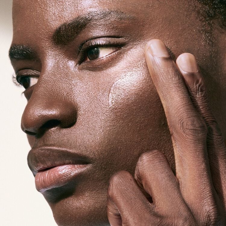 A person with deep brown skin and short dark hair applies a clear, glossy product to their cheek with their fingertips, looking off to the side with a serious expression.