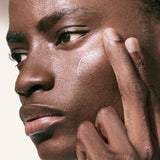 A person with deep brown skin and short dark hair applies a clear, glossy product to their cheek with their fingertips, looking off to the side with a serious expression.