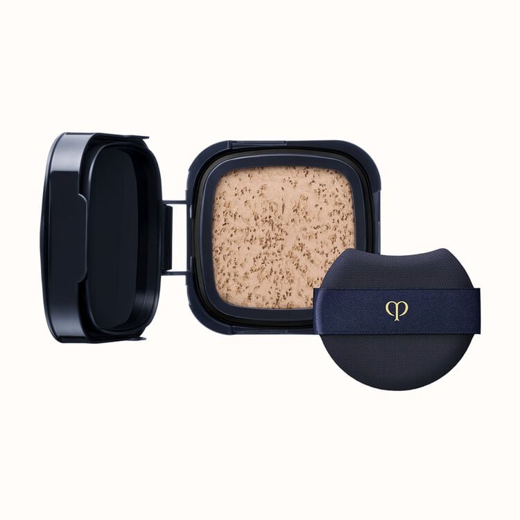 A black compact with an open lid revealing light-colored pressed powder, next to a black cushion puff with a gold logo on a dark blue ribbon.