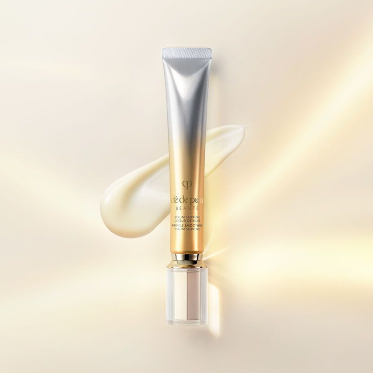 A tube of Clé de Peau Beauté face serum with a silver top and gold bottom, next to a dollop of the product on a white and gold background.